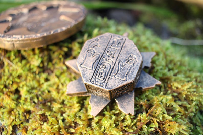 May include: Bronze-colored, octagonal pendant with hieroglyphic carvings and a star-shaped base. Another bronze coin is visible in the background, all resting on a bed of green moss. The pendant has a detailed, aged appearance.