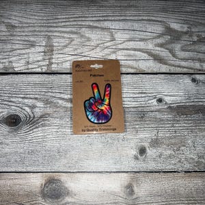 Könnte beinhalten: Ein Peace-Zeichen-Patch im Tie-Dye-Stil mit einem Regenbogen-Farbmuster. Der Patch ist 10 cm x 10 cm groß und besteht aus 50 % Rayon und 50 % Polyester. Der Patch ist mit "Patches" und "by Quality Trimmings" beschriftet.