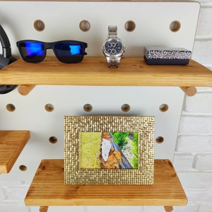 May include: A white pegboard with two wooden shelves. The top shelf has a pair of blue sunglasses, a silver watch, and a small black box. The bottom shelf has a gold picture frame with a photo of a white cat.