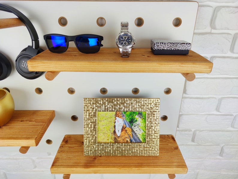 May include: A white pegboard with two wooden shelves. The top shelf has a pair of blue sunglasses, a silver watch, and a small black box. The bottom shelf has a gold picture frame with a photo of a white cat.