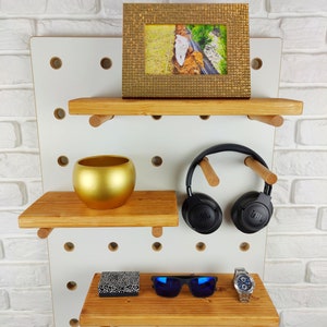 May include: White pegboard with three wooden shelves. The top shelf has a gold picture frame with a photo of a white cat. The middle shelf has a gold planter. The bottom shelf has a small black box, blue sunglasses, and a silver watch.