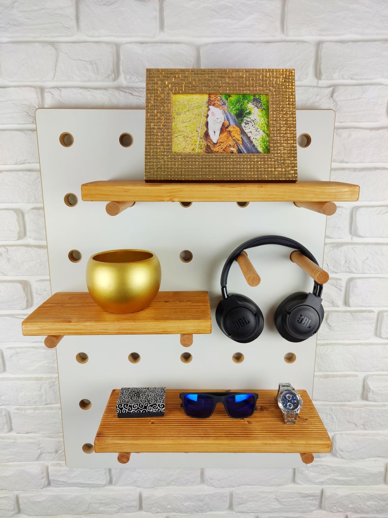 May include: White pegboard with three wooden shelves. The top shelf has a gold picture frame with a photo of a white cat. The middle shelf has a gold planter. The bottom shelf has a small black box, blue sunglasses, and a silver watch.