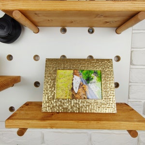 May include: A gold picture frame with a textured surface sits on a wooden shelf. The frame holds a photo of a white cat walking on a path.