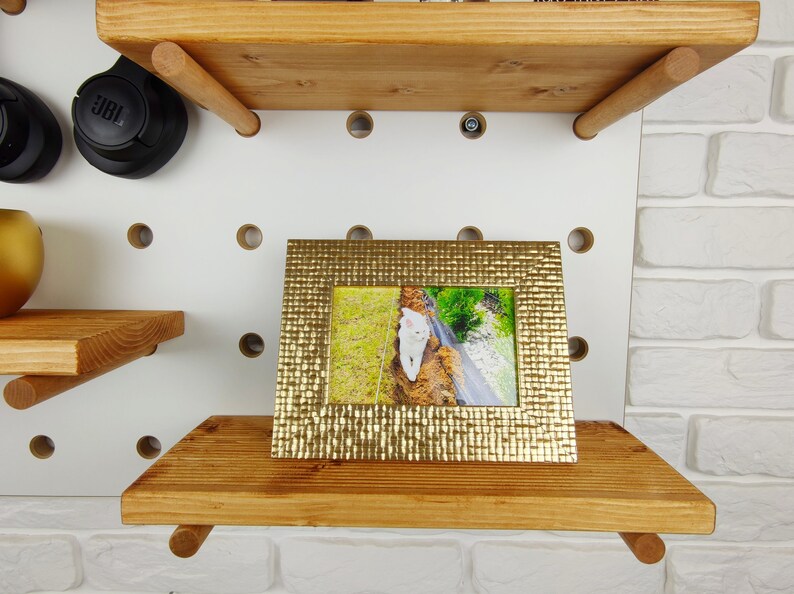 May include: A gold picture frame with a textured surface sits on a wooden shelf. The frame holds a photo of a white cat walking on a path.