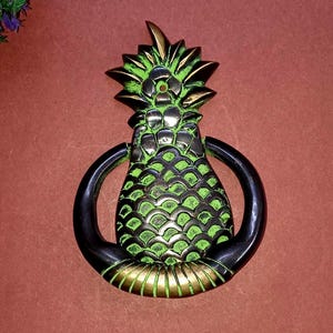 Op de afbeelding: Een decoratieve deurklopper in de vorm van een ananas met een textuurrijke groene en bronzen afwerking. Het centrale ananas-motief is omgeven door een gladde, donkere, gebogen metalen ring die dient als het slagdeel voor het deurbeslag.