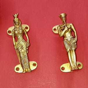 May include: Two gold-coloured brass door handles shaped like standing Egyptian figures. Each handle features a mounting plate at the top and bottom with screw holes. The figures are depicted in traditional attire with detailed carvings.