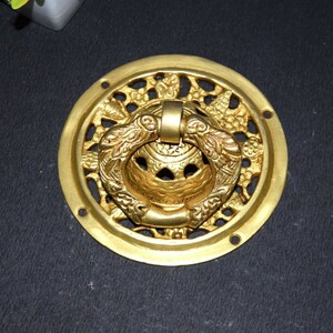 Puede incluir: Un llamador de puerta de metal dorado con un diseño intrincado. El llamador es redondo y tiene un asa de anillo. El diseño presenta un patrón floral y un anillo central.