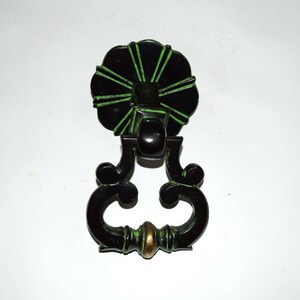 May include: A black and green decorative drawer pull with a floral design. The pull has a circular top with green accents, a central knob, and a curved handle with a brass-colored accent. The hardware is likely made of metal.