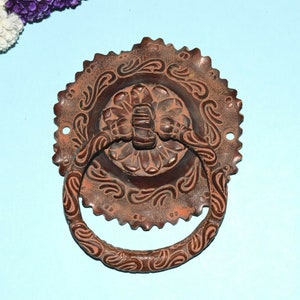 Puede incluir: Un llamador de puerta redondo de cobre, decorativo y adornado, con un asa en forma de anillo. El llamador tiene un diseño floral detallado.