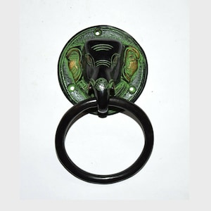 Puede incluir: Un llamador de puerta de metal negro y verde con un diseño de cabeza de elefante. Las orejas del elefante están acentuadas con pintura dorada.
