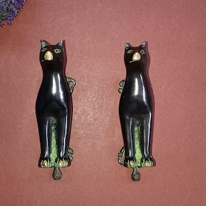 Könnte beinhalten: Zwei passende Türklopfer aus Metall in Form einer schwarzen Katze mit messingfarbenen Akzenten an Ohren, Nase, Augen und Pfoten. Jede Katzenfigur hat ein vertikales Design mit einem kleinen runden Montageloch an der Unterseite zur Befestigung.