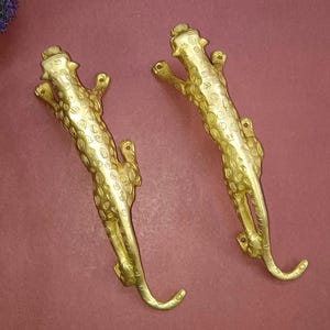 Puede incluir: Dos manijas de puerta de metal dorado con forma de leopardos saltando. Cada manija presenta un patrón texturizado y moteado en todo el cuerpo, con orificios de montaje visibles en la cabeza y la cola para una fijación segura a una puerta o mueble.