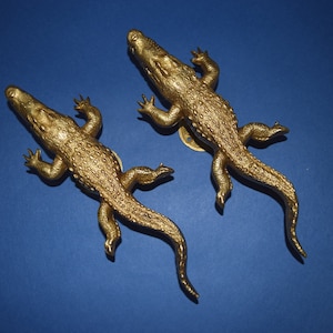 May include: Two gold-colored alligator door handles. The handles are detailed with textured scales and have a realistic appearance.