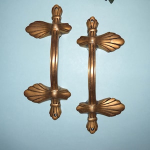 May include: A pair of antique gold cabinet handles. Each handle has a vertical bar with decorative top and bottom sections, and a central grip. The design incorporates a fan-shaped element at each end, providing an elegant touch.