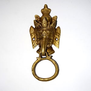 May include: A gold-toned door knocker shaped like the head of an elephant with a crown. The knocker has a ring for pulling.
