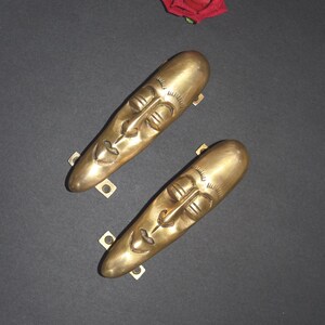 May include: Two gold-toned door handles shaped like stylised faces with closed eyes. The handles are mounted on a black surface.