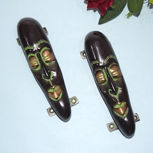 Könnte beinhalten: Zwei schwarze und grüne Türgriffe in Form einer Gesichtsmaske mit goldenen Akzenten. Die Griffe sind mit Metallhalterungen an einer Wand befestigt.