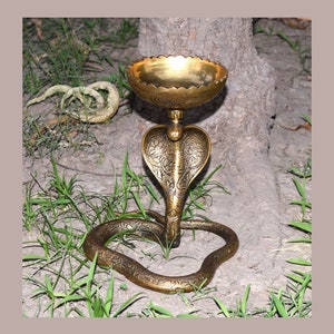 Puede incluir: Un quemador de incienso de metal dorado con forma de cobra y un cuenco decorativo en la parte superior. El quemador está sentado sobre un lecho de hierba verde.