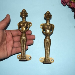 May include: Two brass door handles in the shape of standing women. The handles are detailed with intricate designs and have a gold finish.