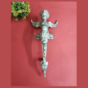 Peut inclure: Poignée de porte en métal argenté en forme d'ange avec les ailes déployées. L'ange a un visage chérubin et tient une fleur dans chaque main. La poignée est ornée et détaillée.