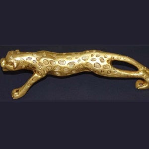Puede incluir: Una figura de metal de color dorado que representa a un leopardo en posición de acecho. La escultura presenta una superficie texturizada con manchas en relieve por todo el cuerpo, una cola larga que se curva hacia arriba y cabeza detallada.