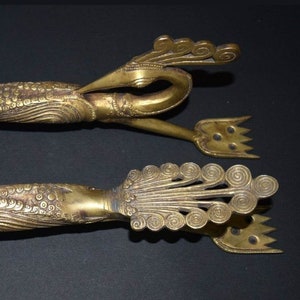 Może przedstawiać: Dwa ozdobne mosiężne klamki do drzwi w kształcie pawia z misternymi detalami. Klamki są w stylu vintage i mają złote wykończenie.