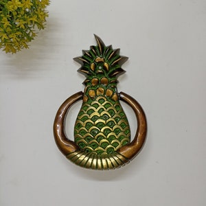 May include: A brass pineapple door knocker with a green and gold finish. The knocker is in the shape of a pineapple with a ring around the top for knocking.