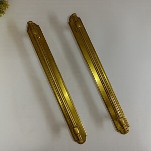 May include: Two gold-toned metal door handles with a decorative design. The handles are long and narrow with a slightly curved shape.