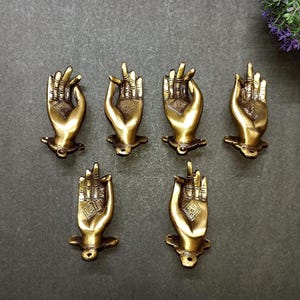 May include: Six antique brass-colored hand-shaped decorative objects. Each hand is in a different gesture, with intricate details on the fingers and palm. The hands are mounted on a small base, and are arranged in a circular pattern.