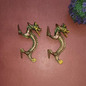 May include: A pair of antique brass dragon-shaped wall hooks. The hooks have green and gold detailing, with a curved body and a dragon head. They are set against a reddish-brown background, with a small purple floral arrangement visible.