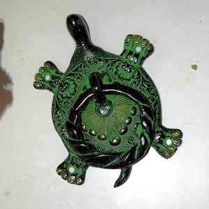 May include: A turtle-shaped door knocker made of dark metal with a green finish. The shell features intricate floral scrollwork and a central circular ring with a twisted rope texture. The head, legs, and tail are detailed with gold-toned accents.
