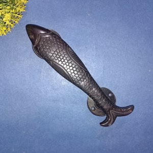 Op de afbeelding: Een zwarte metalen deurknop in de vorm van een vis met een getextureerd oppervlak. De knop is ontworpen om eruit te zien als een vis met schubben en vinnen.