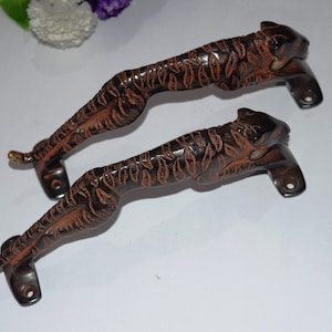 Könnte beinhalten: Zwei verzierte Türgriffe in Form von hockenden Tigern. Die Griffe sind aus dunkelbraunem Metall mit einer strukturierten Oberfläche und goldenen Akzenten.