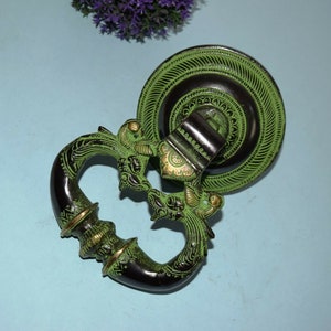 Może przedstawiać: Mosiężny klamka do drzwi w kolorze zielonym i czarnym z ozdobnym wzorem. Klamka ma kształt dwóch ptaków z głowami zwróconymi do siebie. Klamka jest przymocowana do okrągłej metalowej płytki z ozdobną ramką.