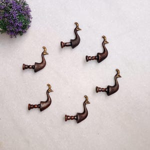 Könnte beinhalten: Sechs bronzefarbene Gardinenhalter in Pfauenform mit goldenen Akzenten. Die Halter sind auf einer weißen Oberfläche angeordnet, mit einem kleinen Strauß lila Kunstblumen. Der Kopf des Pfaus ist nach oben gerichtet.