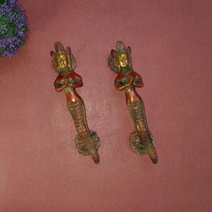 以下が含まれることがあります： 祈りを捧げる女性の形をした、装飾的な真鍮製のドアノブが2つ。ノブは赤みがかった茶色で、金色のアクセントが施されています。