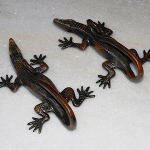May include: Two bronze lizard figurines with curled tails. The lizards are facing opposite directions and have detailed scales and claws.