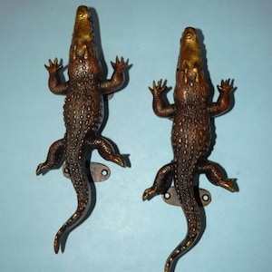 May include: A pair of decorative door handles shaped like crocodiles. The handles are made of metal and have a bronze finish. The crocodiles are in a standing position with their mouths open.