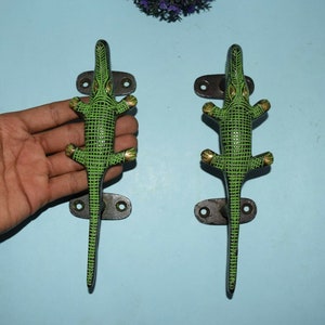 May include: A pair of green and gold crocodile door handles with black metal bases. The handles are designed to look like a crocodile with its mouth open.