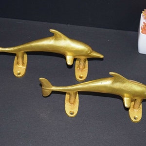 Op de afbeelding: Twee gouden dolfijn deurgrepen. De grepen zijn gemaakt van metaal en hebben een realistisch dolfijn ontwerp. Ze zijn gemonteerd op een muur.