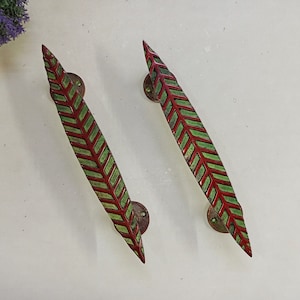 Może przedstawiać: Dwa zielono-czerwone uchwyty do drzwi w kształcie liści, z mosiężnym wykończeniem.