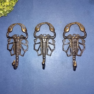 May include: Three gold-toned metal scorpion hooks. The hooks are in the shape of a scorpion with a curled tail. The hooks are on a blue background.