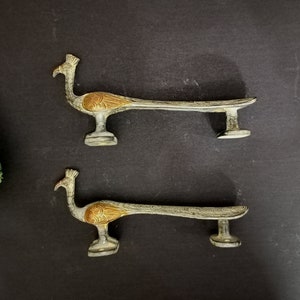 May include: Two silver peacock door handles with gold accents. The handles are shaped like peacocks with their tails spread out. They are mounted on a black background.