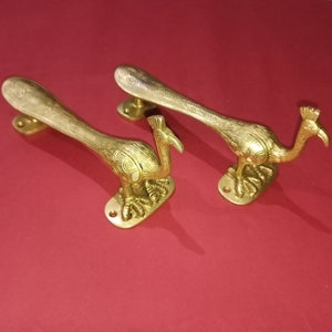 Op de afbeelding: Twee gouden pauw deurgrepen met ingewikkelde details. De grepen zijn gevormd als pauwen met hun kop naar de zijkant gedraaid. De grepen zijn gemonteerd op een vierkante basis.