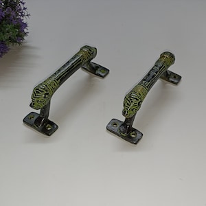 Può includere: Due maniglie per porte in metallo nero e verde con dettagli decorativi. Ogni maniglia ha un design a testa di leone a un'estremità e una base rettangolare con due fori per viti.