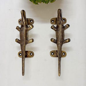 May include: A pair of bronze-toned alligator-shaped door handles. Each handle has a textured scale pattern and a mounting plate with two screw holes. The handles are set against a white backdrop.