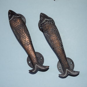 Puede incluir: Dos tiradores de puerta de pescado de estilo antiguo con un acabado en bronce oscuro. Los tiradores están detallados con escamas y aletas.