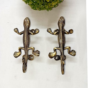Peut inclure: Deux crochets décoratifs en forme de lézard de couleur bronze. Les lézards présentent des écailles texturées et des membres tendus, conçus pour être fixés au mur. Idéal pour une décoration rustique ou sur le thème de la nature.