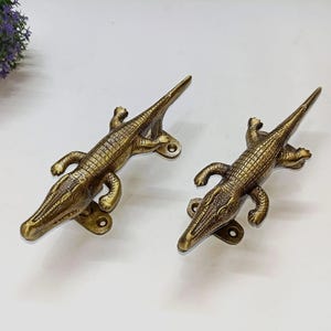 May include: Two antique brass-toned metal door handles shaped like crocodiles. Each handle features a textured, scaled body, a long tail, and four legs, with visible mounting holes at the head and tail ends for secure attachment to a door or cabinet surface.