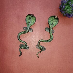 Op de afbeelding: Twee zwarte metalen deurgrepen in de vorm van slangen met groene patronen. De grepen zijn in de vorm van een cobra met zijn kop omhoog.